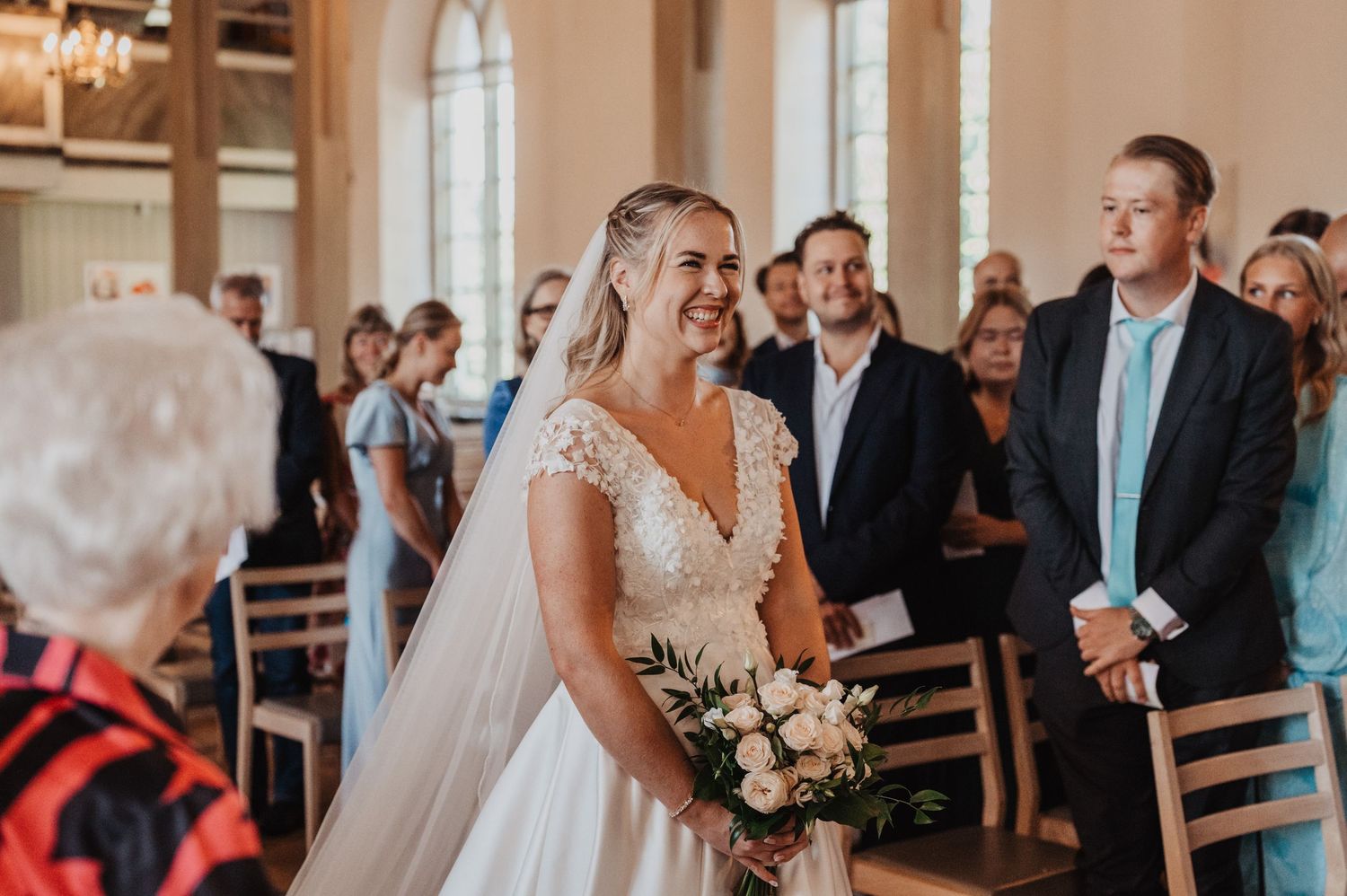 Smiling bride in lace wedding gown holding bouquet walking down the church aisle.