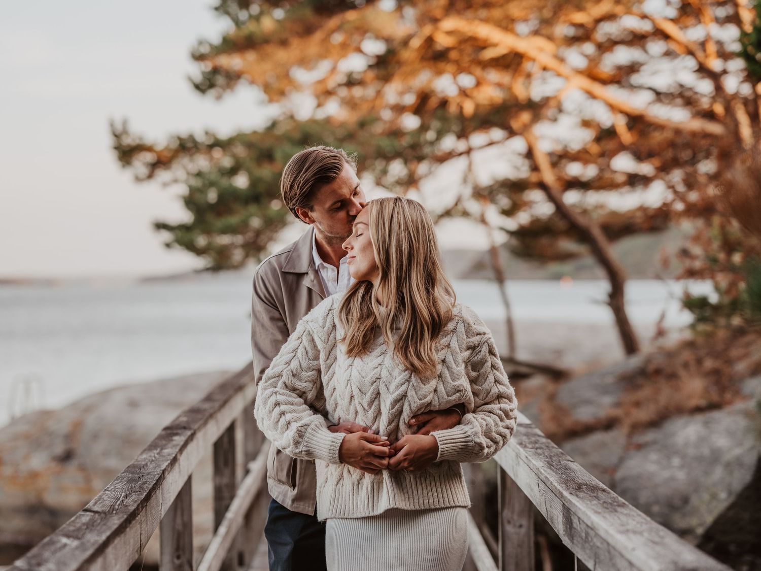 A couple gazes at each other lovingly on a wooden boardwalk near a scenic coastal tree.