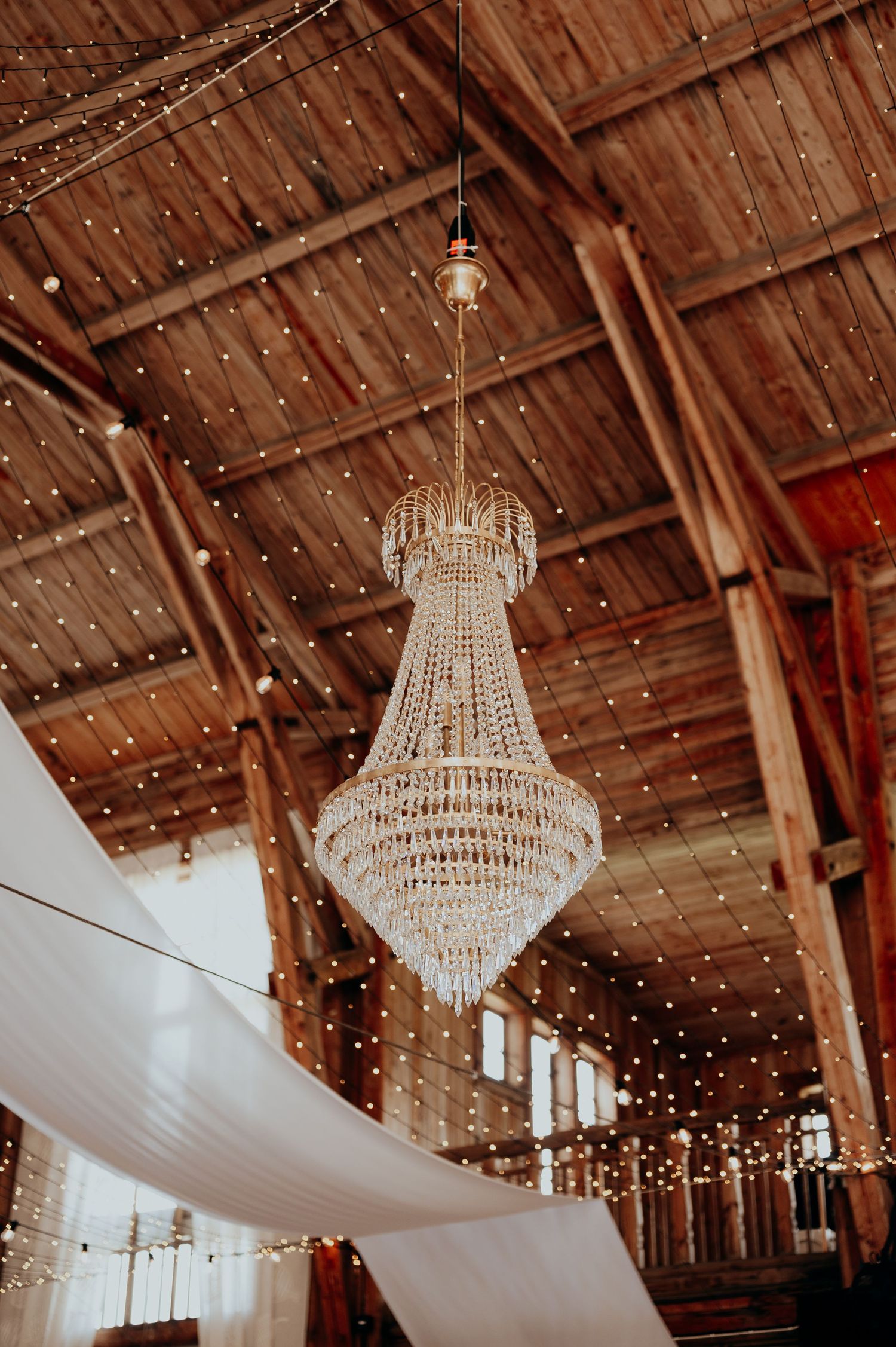 Grand crystal chandelier hanging from wooden beam ceiling with fairy lights in rustic barn venue.