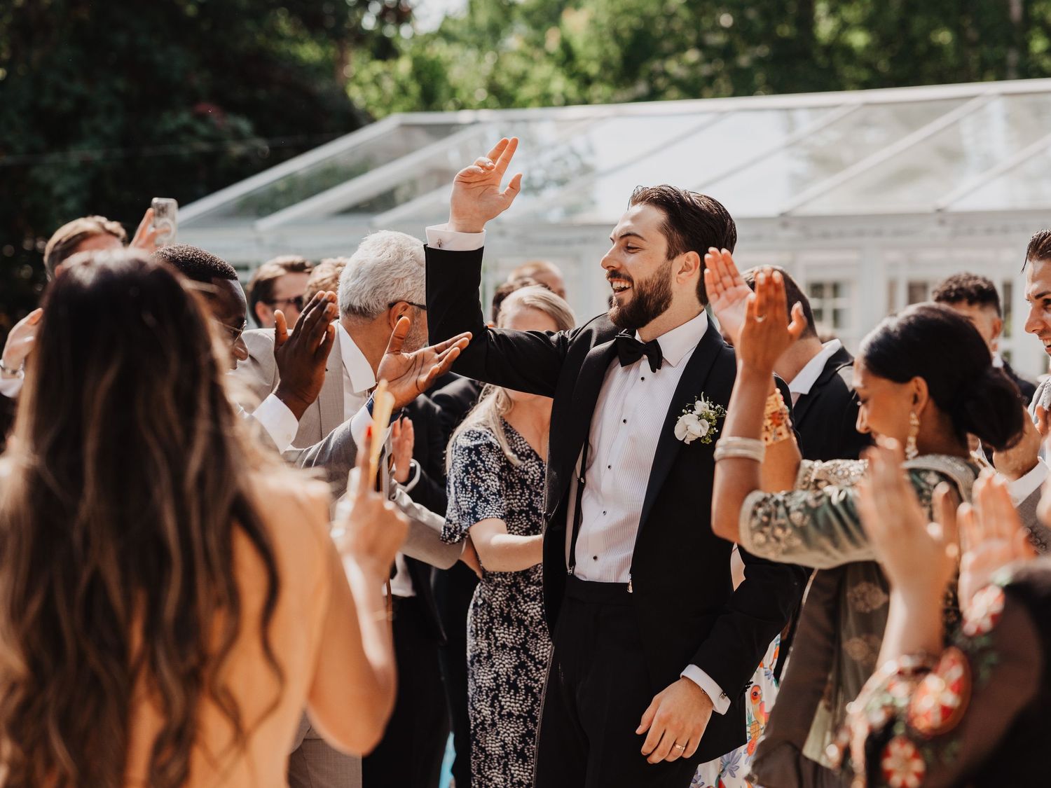 Groom in black tuxedo celebrates with wedding guests outdoors, raising hands joyfully near a glass marquee.
