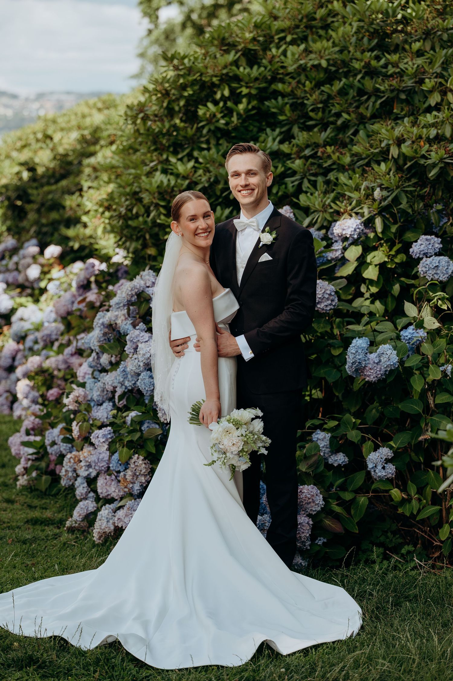 Smiling wedding couple posing before blooming hydrangeas, bride in sleek mermaid gown with long train.