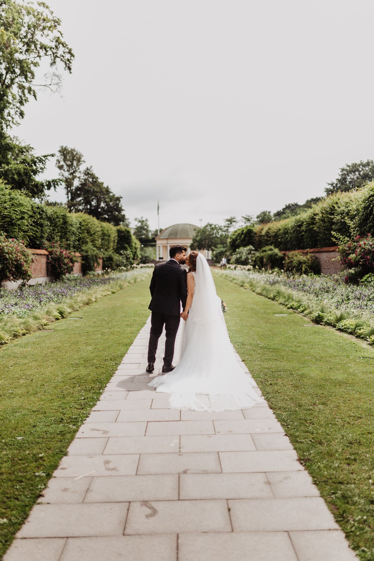 Bride and groom walking hand in hand down a long garden pathway with flowing veil.