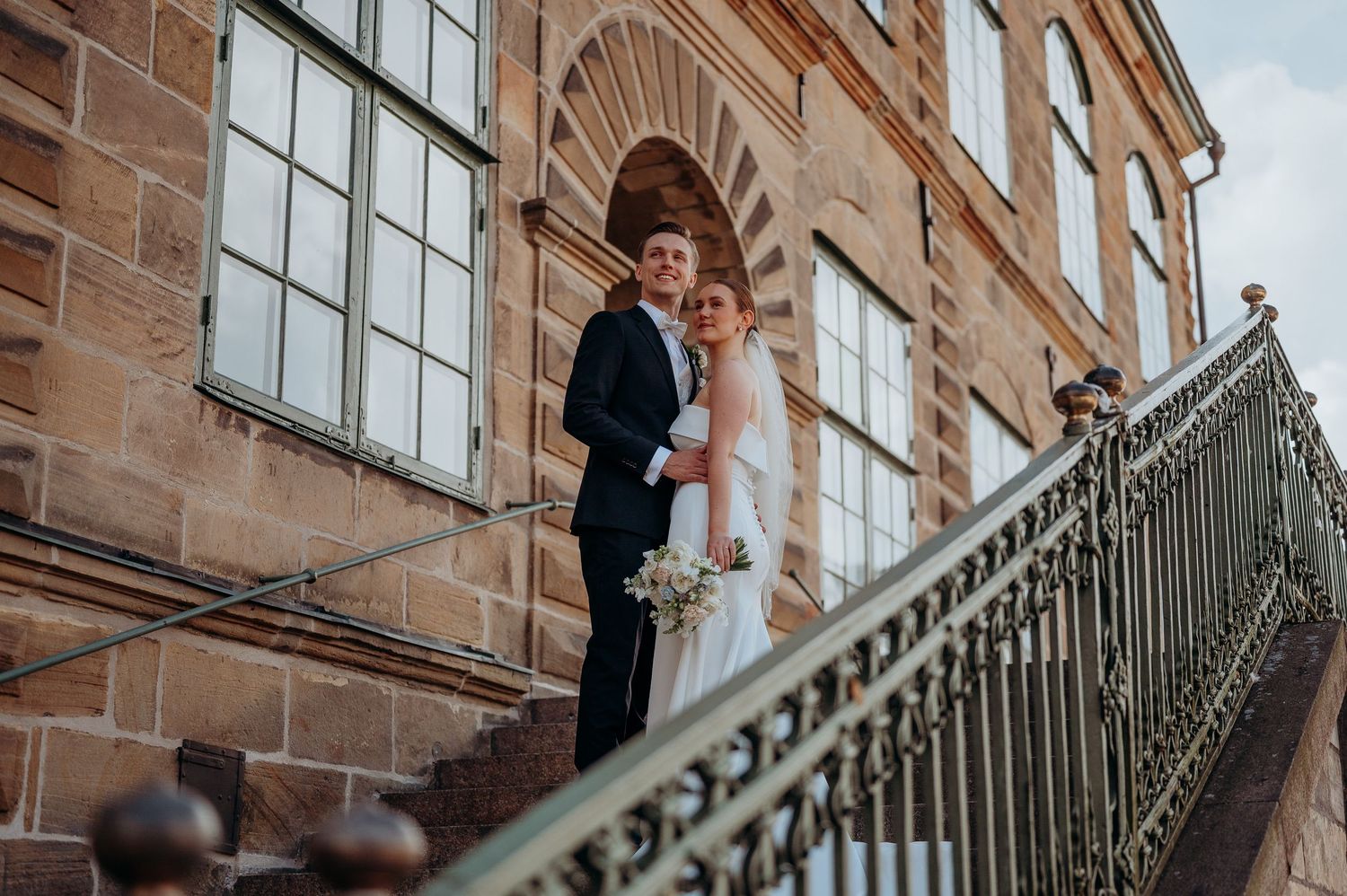 A bride and groom pose on ornate stone steps outside a historic sandstone building on their wedding day.