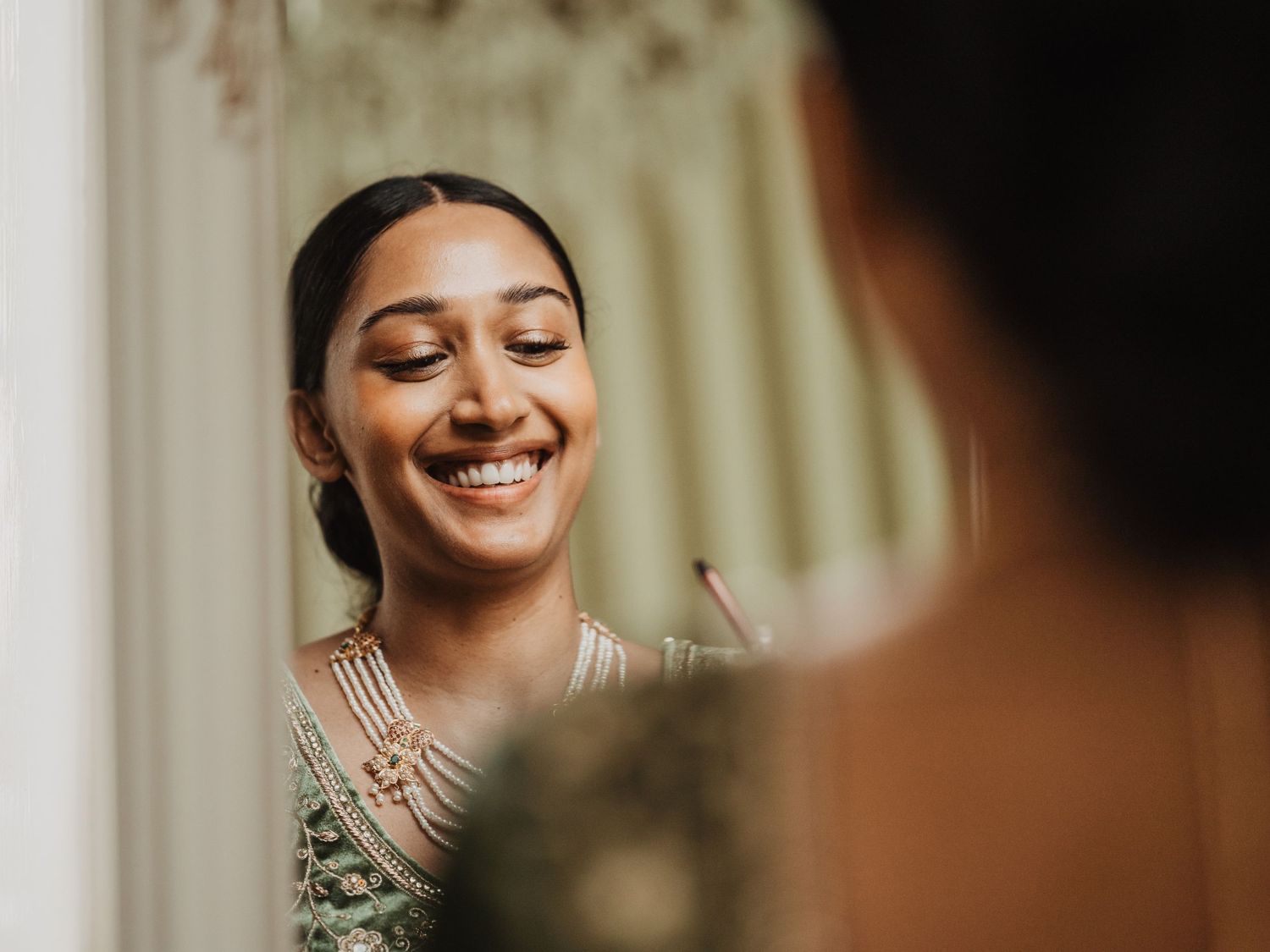 Radiant South Asian bride smiling joyfully at her reflection in the mirror during bridal preparations.