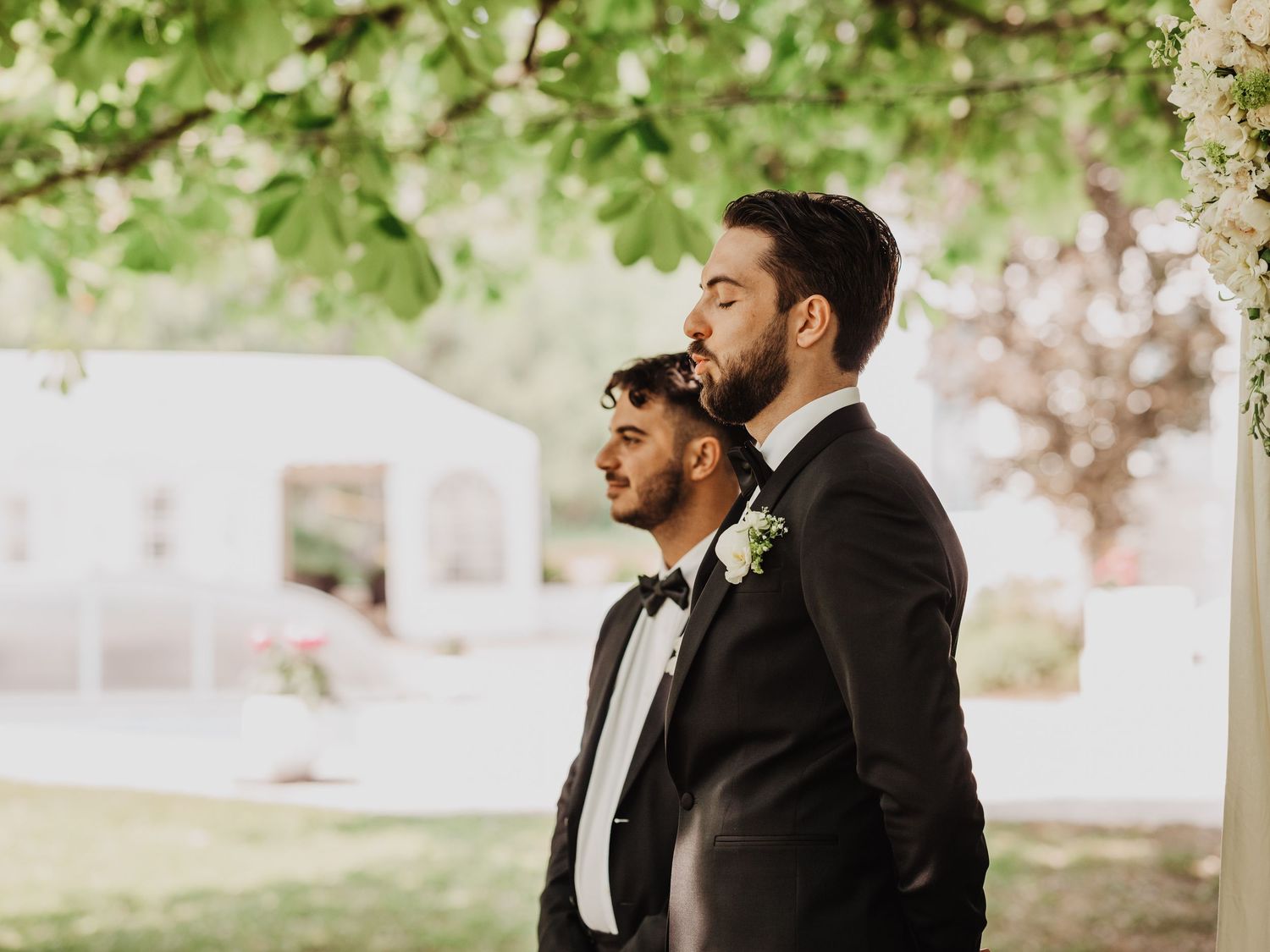 Two grooms in black tuxedos with boutonnieres stand together outdoors under a flowering tree at their wedding.