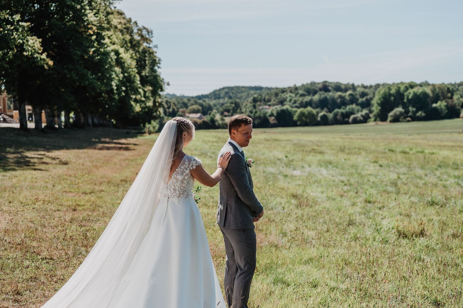 Bride in white gown and veil stands with groom in gray suit in a scenic green field on their wedding day.