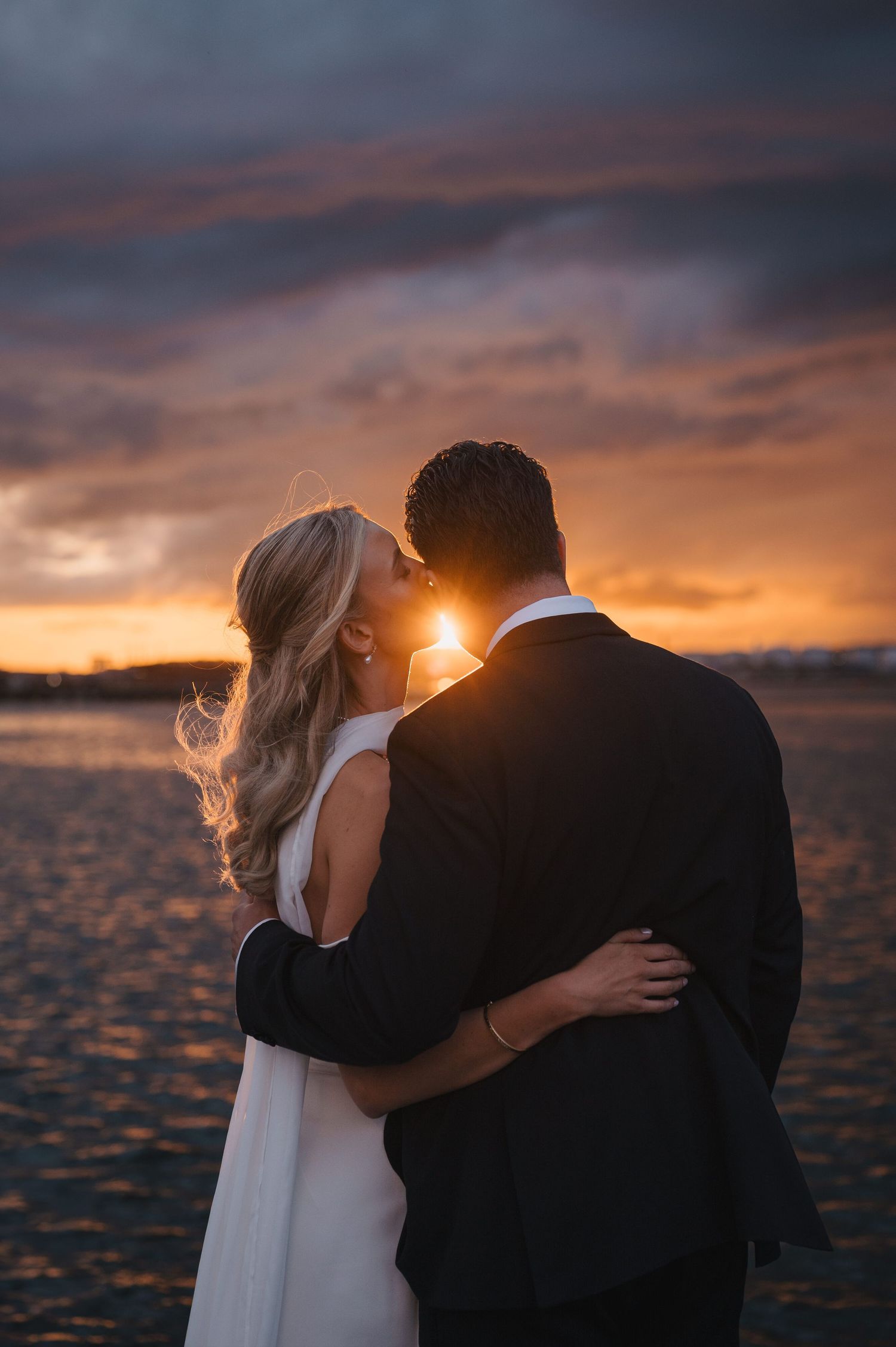 A couple shares a romantic kiss at sunset on a beach, the bride in white and groom in a dark suit.