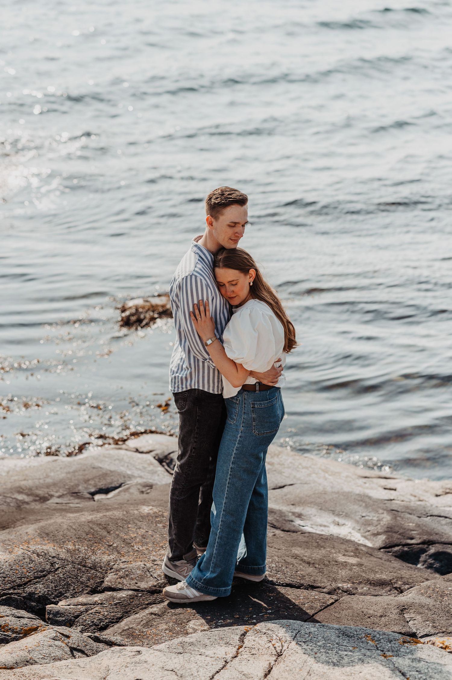 Young couple hugging on rocky shoreline with sparkling ocean water in the background.