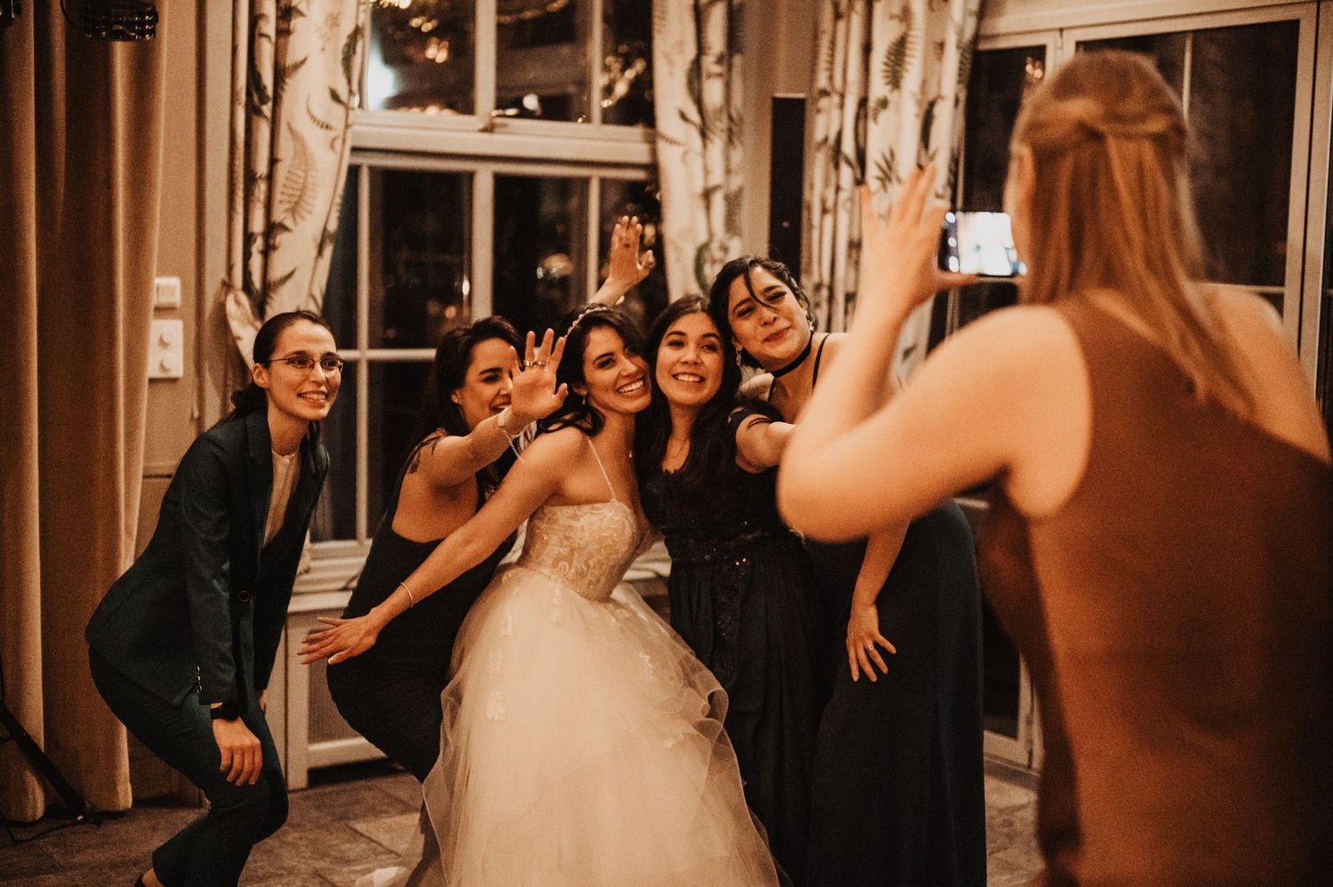 Bride in white gown poses with bridesmaids in navy dresses as friend captures fun group photo at wedding reception.