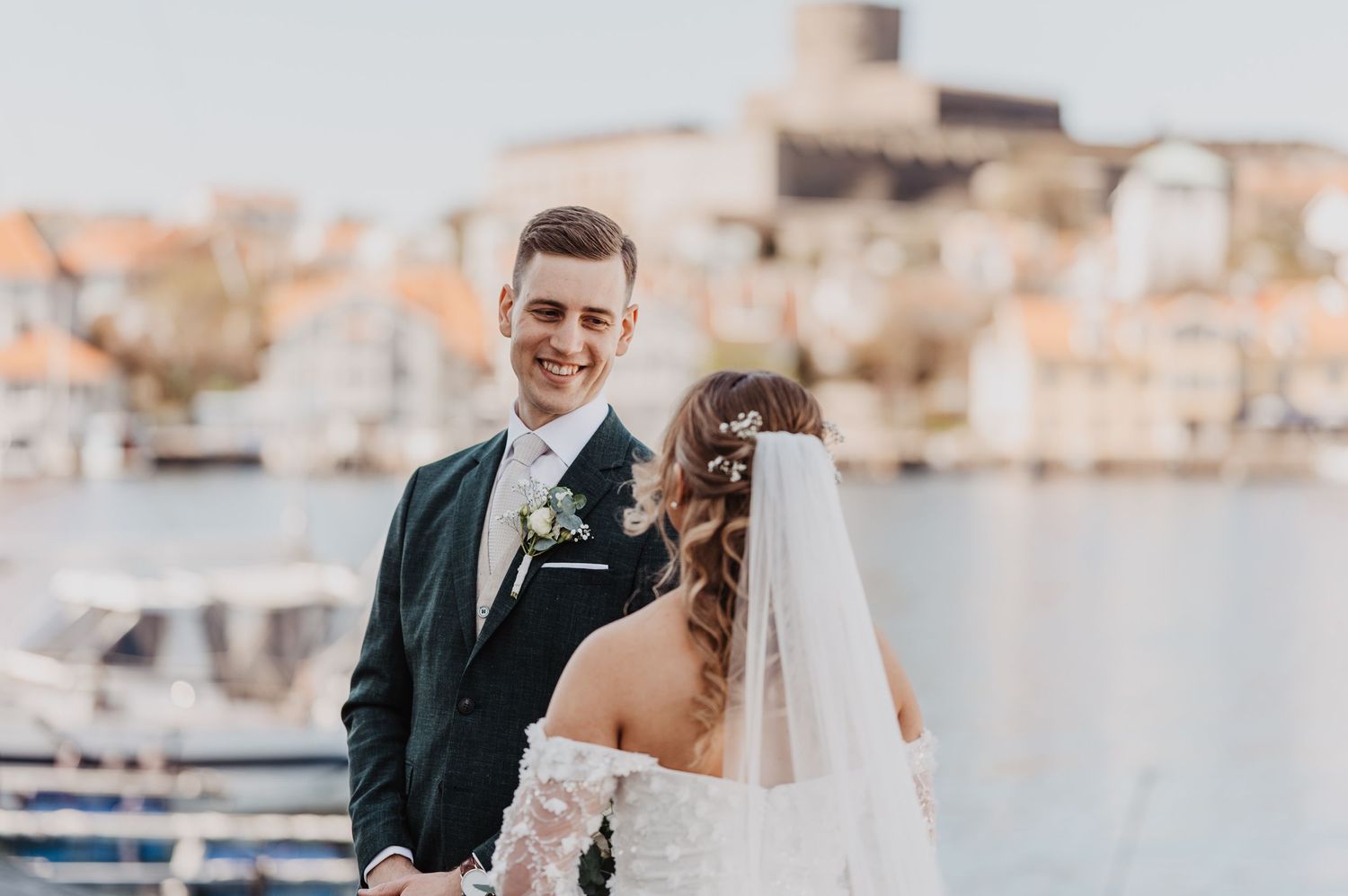 Bride and groom share a romantic moment by the waterfront, with the groom in a dark suit smiling at his veiled bride.