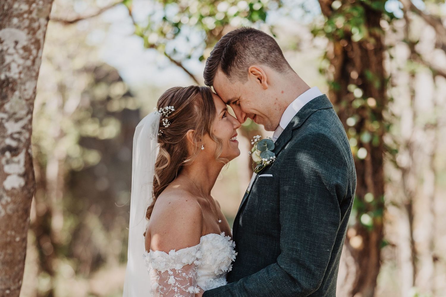 Bride and groom sharing a tender forehead-to-forehead moment surrounded by trees on wedding day.