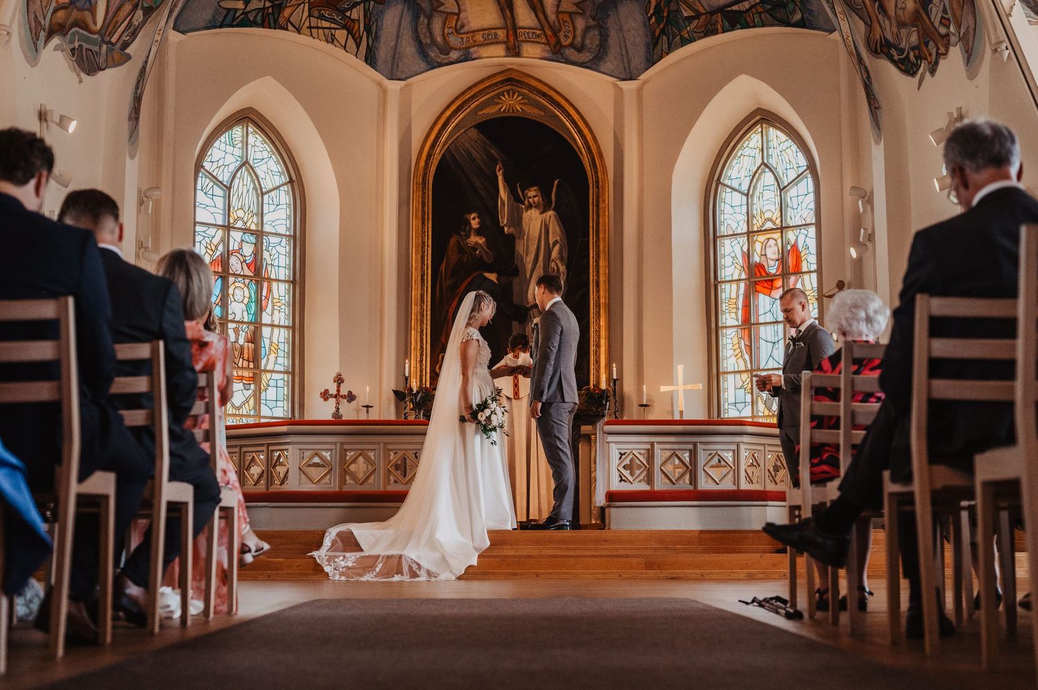 Bride and groom exchange vows at ornate church altar with stained glass windows and wedding guests seated in pews.