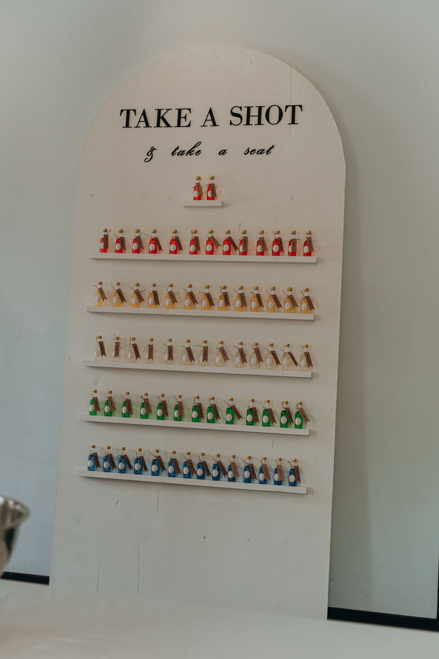 White arched wedding seating display board with colorful mini shot bottles arranged on shelves.