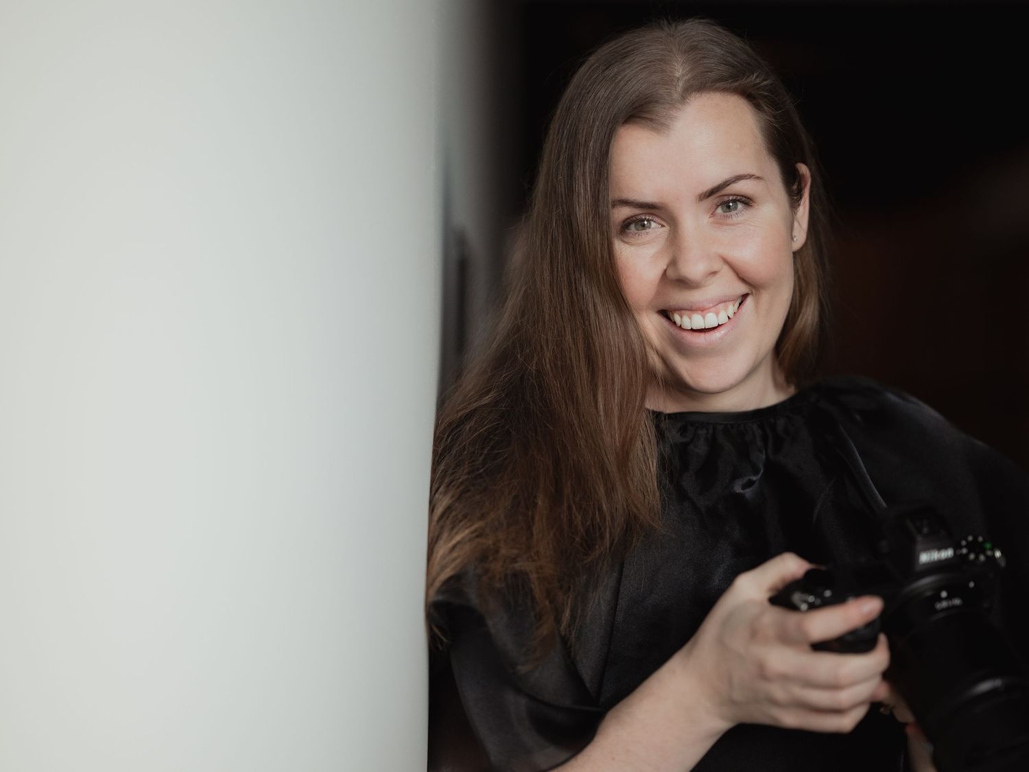 Smiling female photographer holding Nikon camera in black outfit indoors.