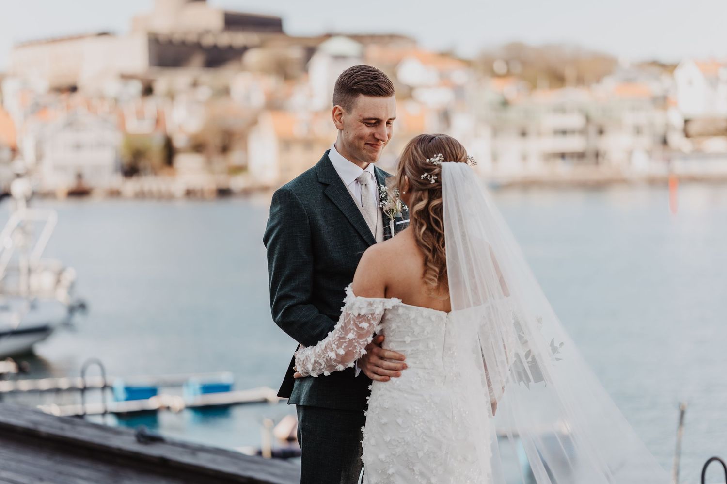 Newlyweds share an intimate gaze on a waterfront dock, bride in lace gown with veil, groom in dark suit.