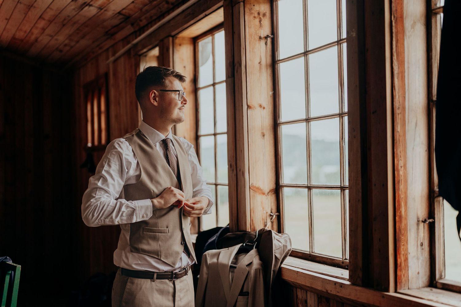 Dapper groom in a grey vest and tie gazing out of a rustic wooden cabin window on his wedding day.