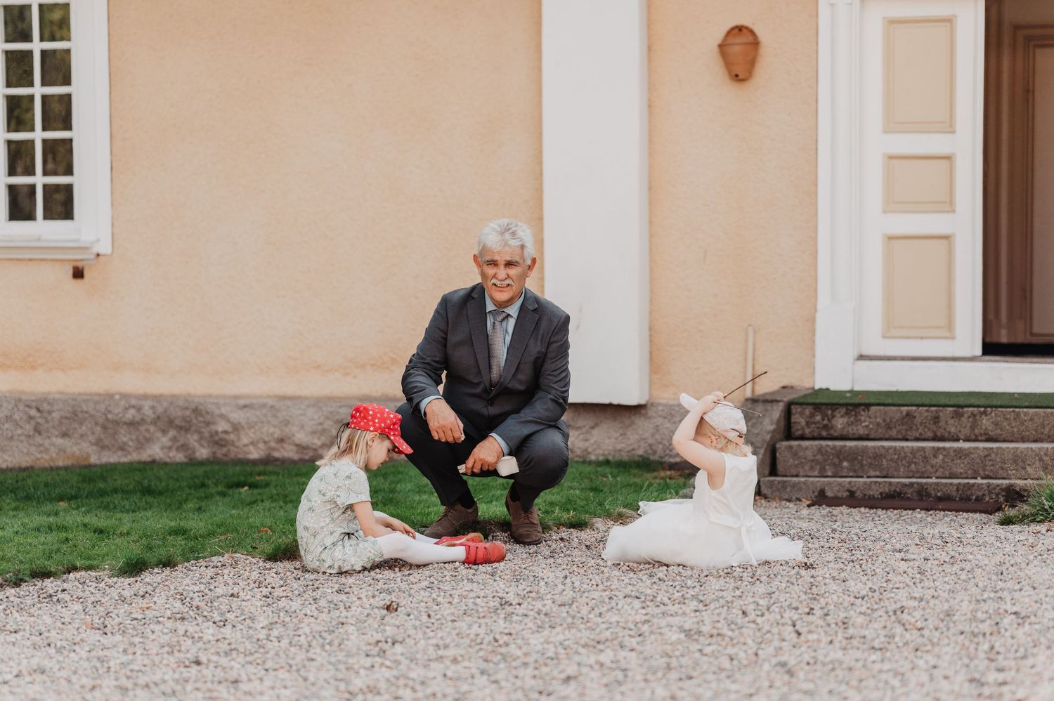 Older man in suit crouches to interact with two small children on gravel path.