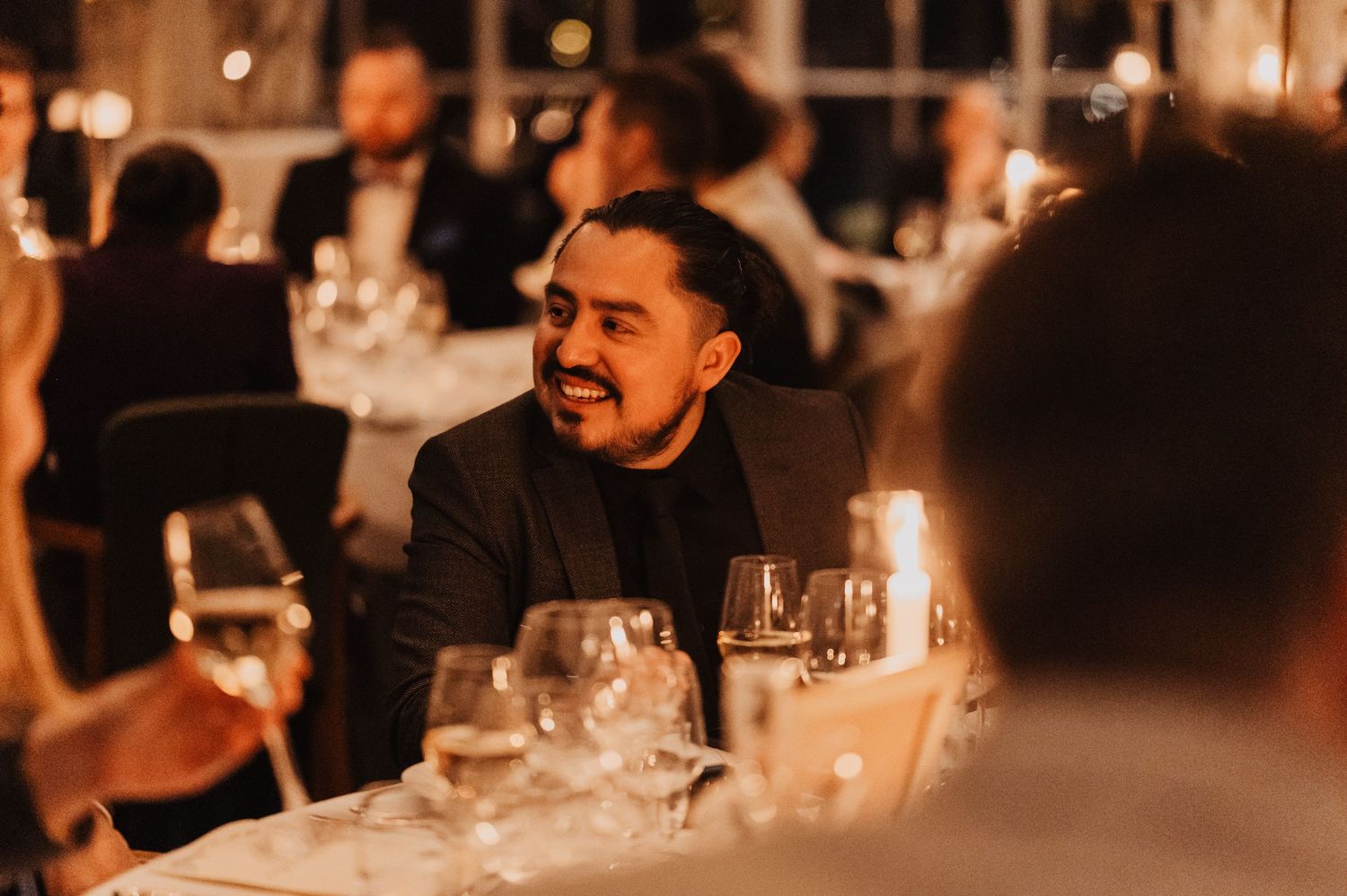 Man in black suit smiling at elegant candlelit dinner party with wine glasses on table.
