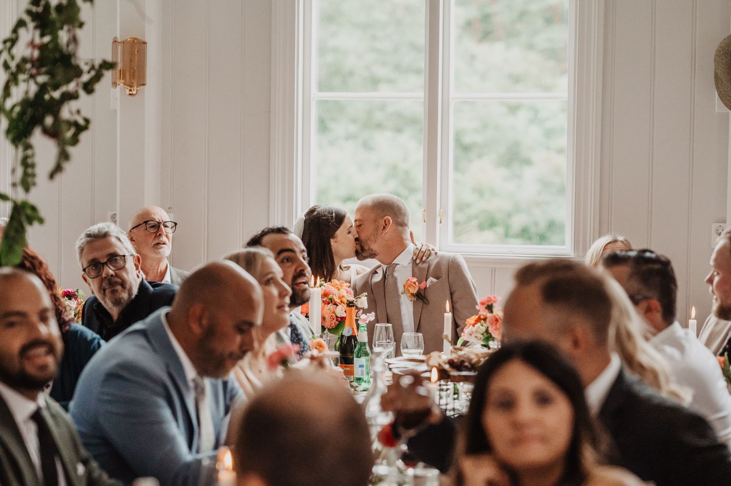 Wedding guests gathered at a long reception table, with the couple sharing a kiss in the background.