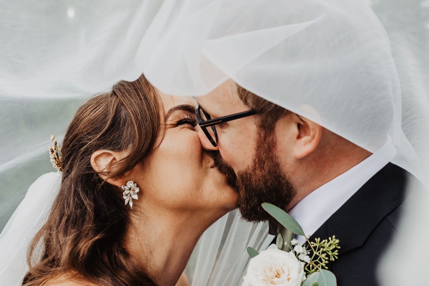 Bride and groom sharing a romantic kiss under a flowing white veil on their wedding day.