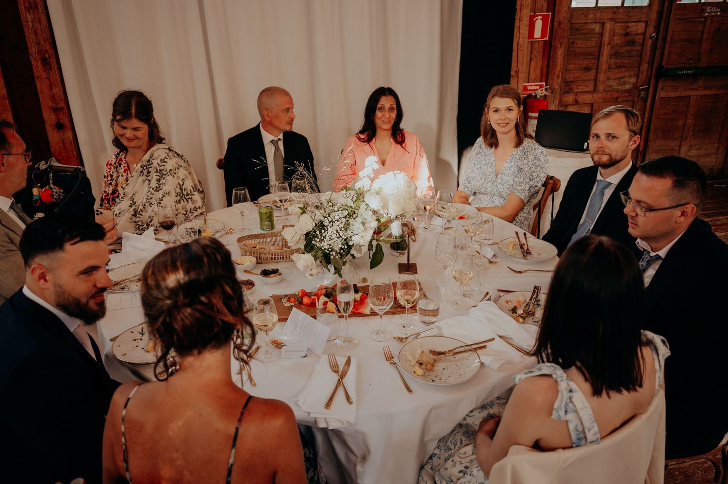 Elegant wedding reception guests seated around a round table with floral centerpiece in a rustic venue.