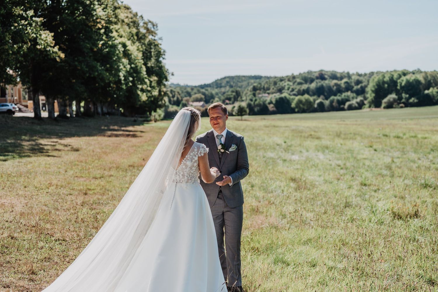 Bride in flowing white gown and veil shares first look moment with groom in gray suit in sunny green field.