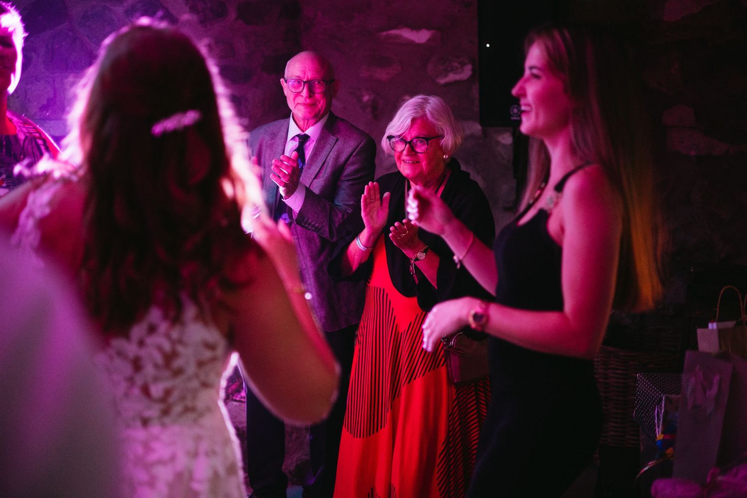 Guests clapping and dancing at a wedding reception with purple lighting in a rustic stone venue.