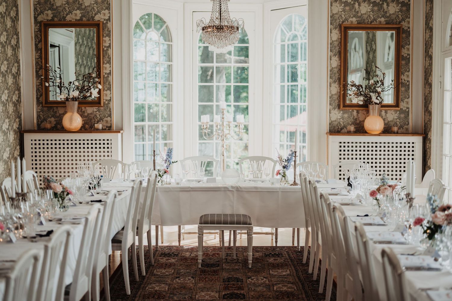 Elegant wedding reception dining room with white tablecloths, floral wallpaper, and chandelier.