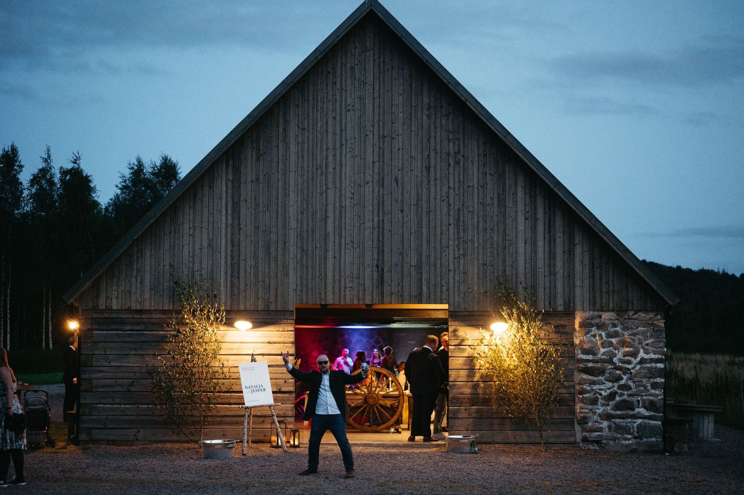Rustic wooden barn wedding venue lit up at dusk with guests gathered at the entrance.