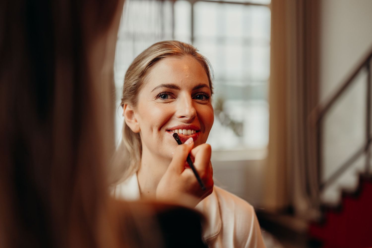 Bride smiling as makeup artist applies red lipstick during wedding preparations.