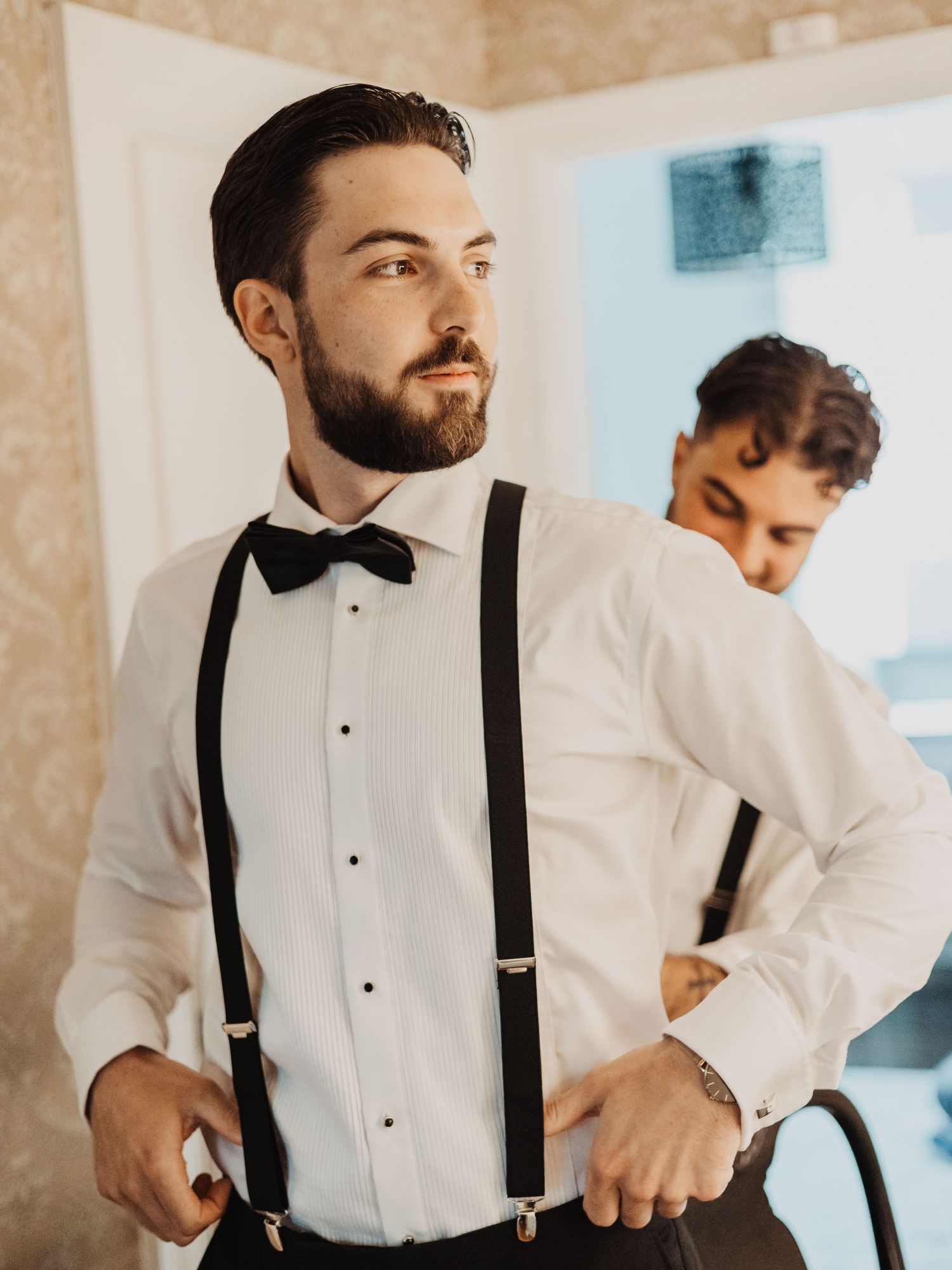 Stylish groom adjusting black suspenders and bow tie while groomsman gets dressed in the background.