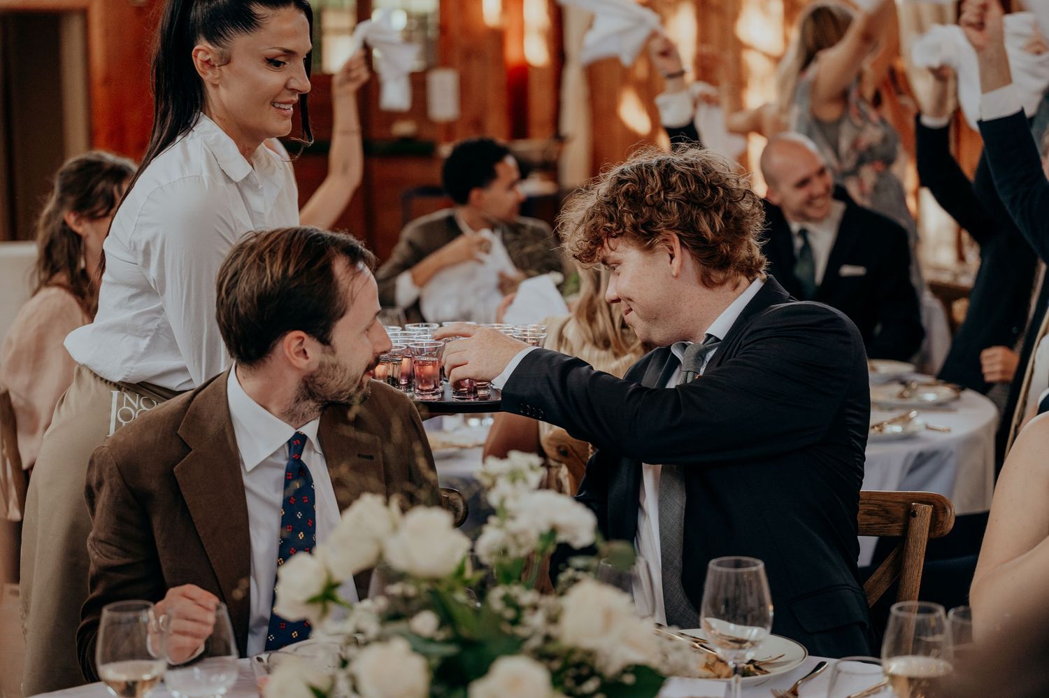 Two men in suits share a handshake at an elegant wedding reception dinner table with floral centerpieces.