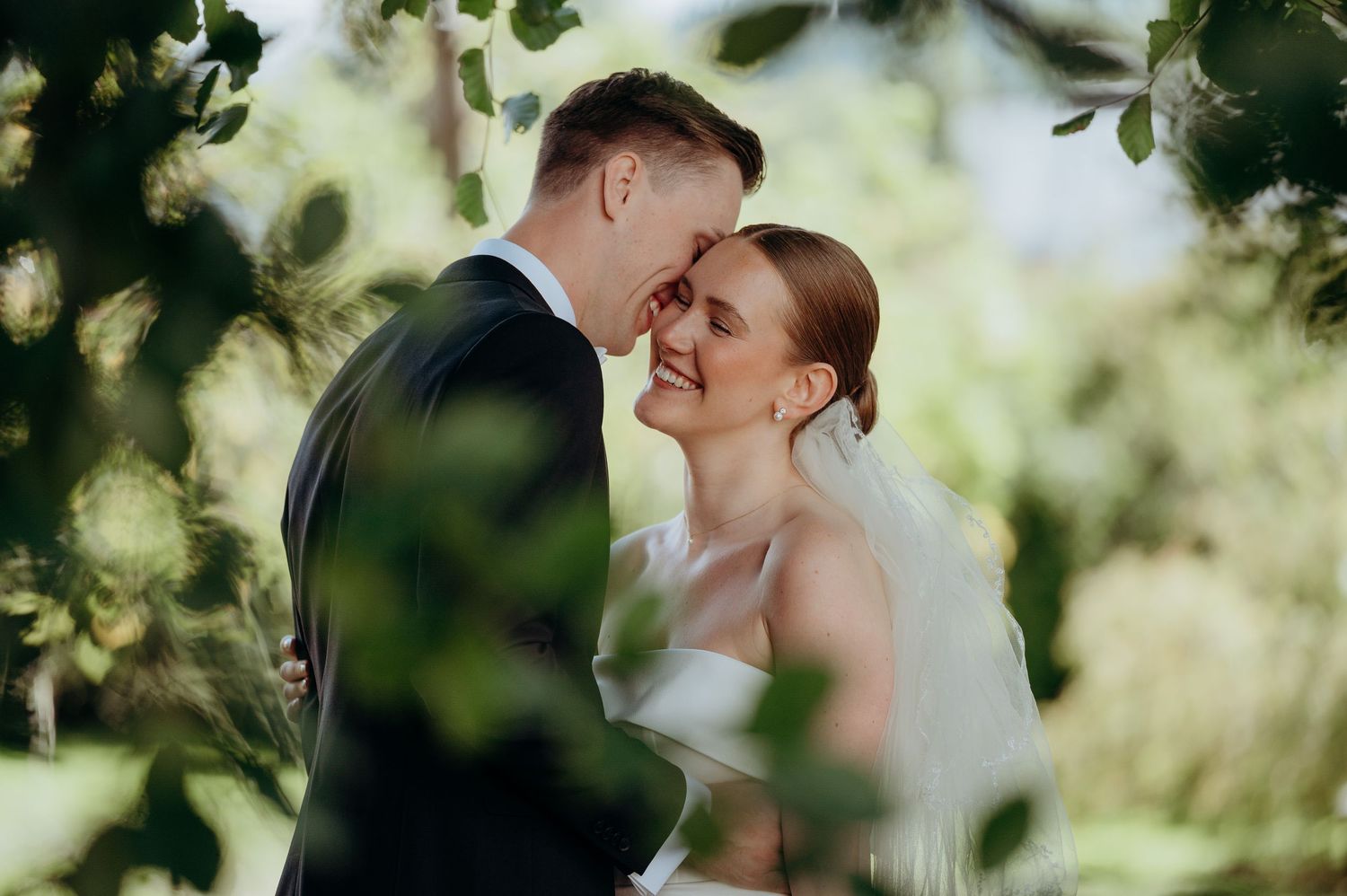 Bride and groom sharing a tender moment surrounded by lush green foliage outdoors.
