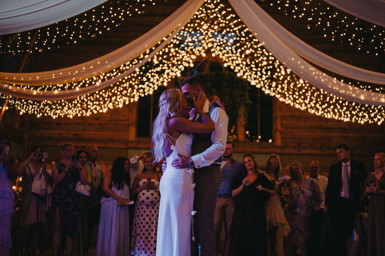Couple shares first dance under fairy lights at romantic barn wedding reception surrounded by guests.