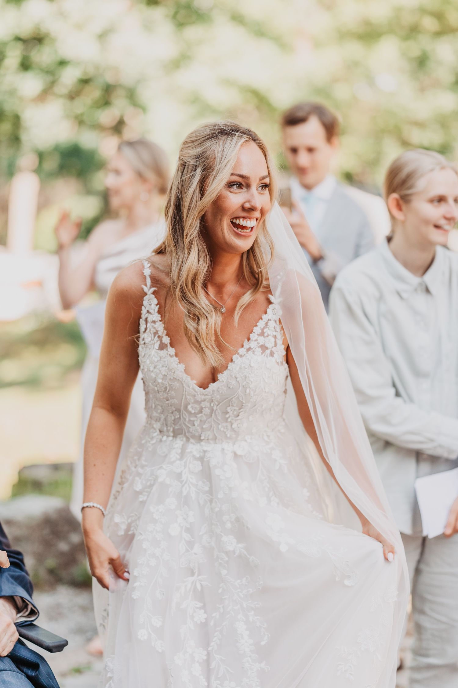 Smiling bride in floral lace wedding gown with veil walks outdoors at garden wedding ceremony.