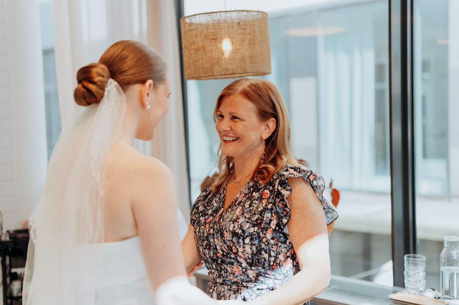 Smiling mother in floral dress shares a warm moment with bride in white gown and veil.