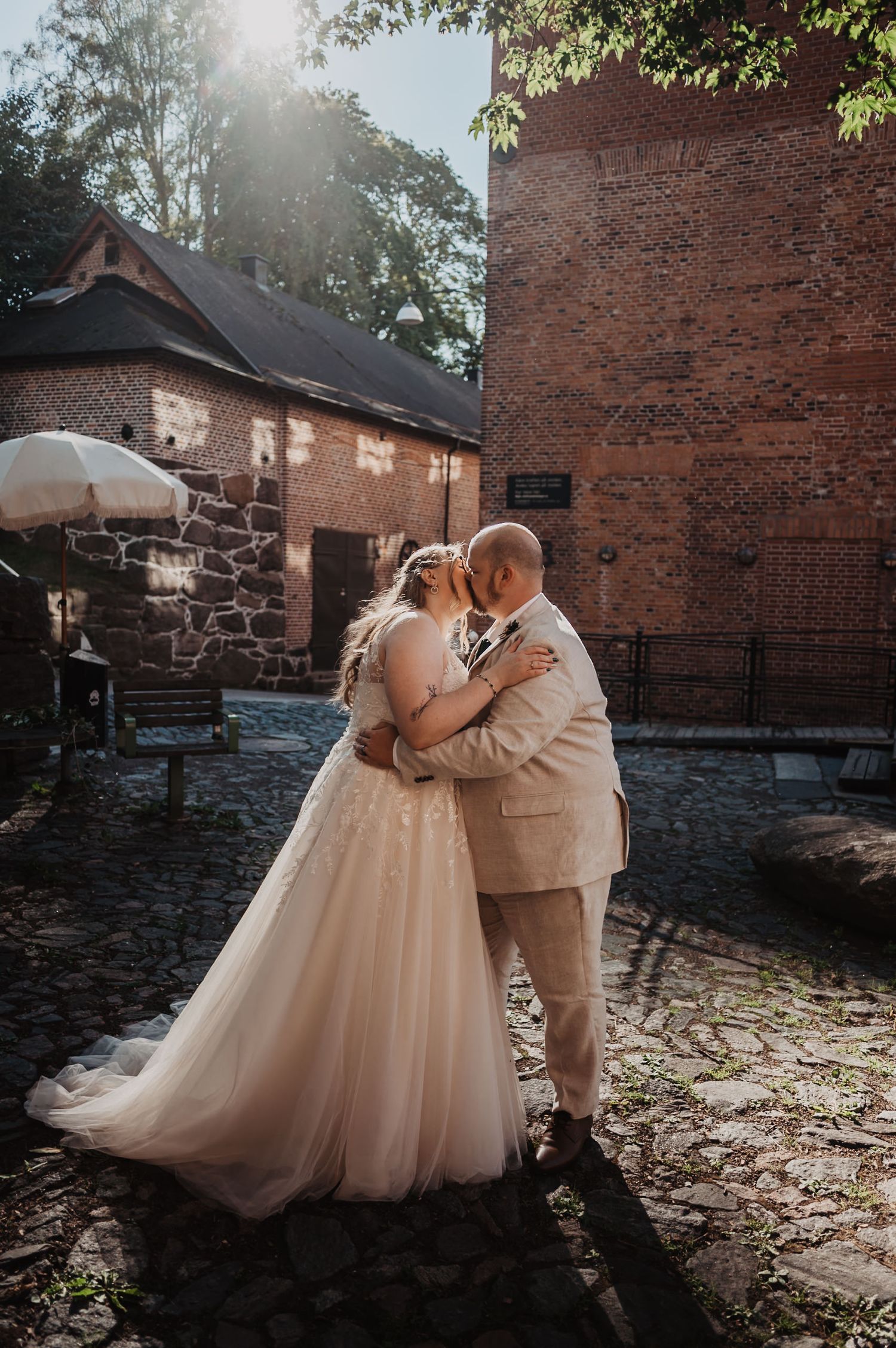 Bride and groom sharing a kiss on cobblestone courtyard beside historic brick building.