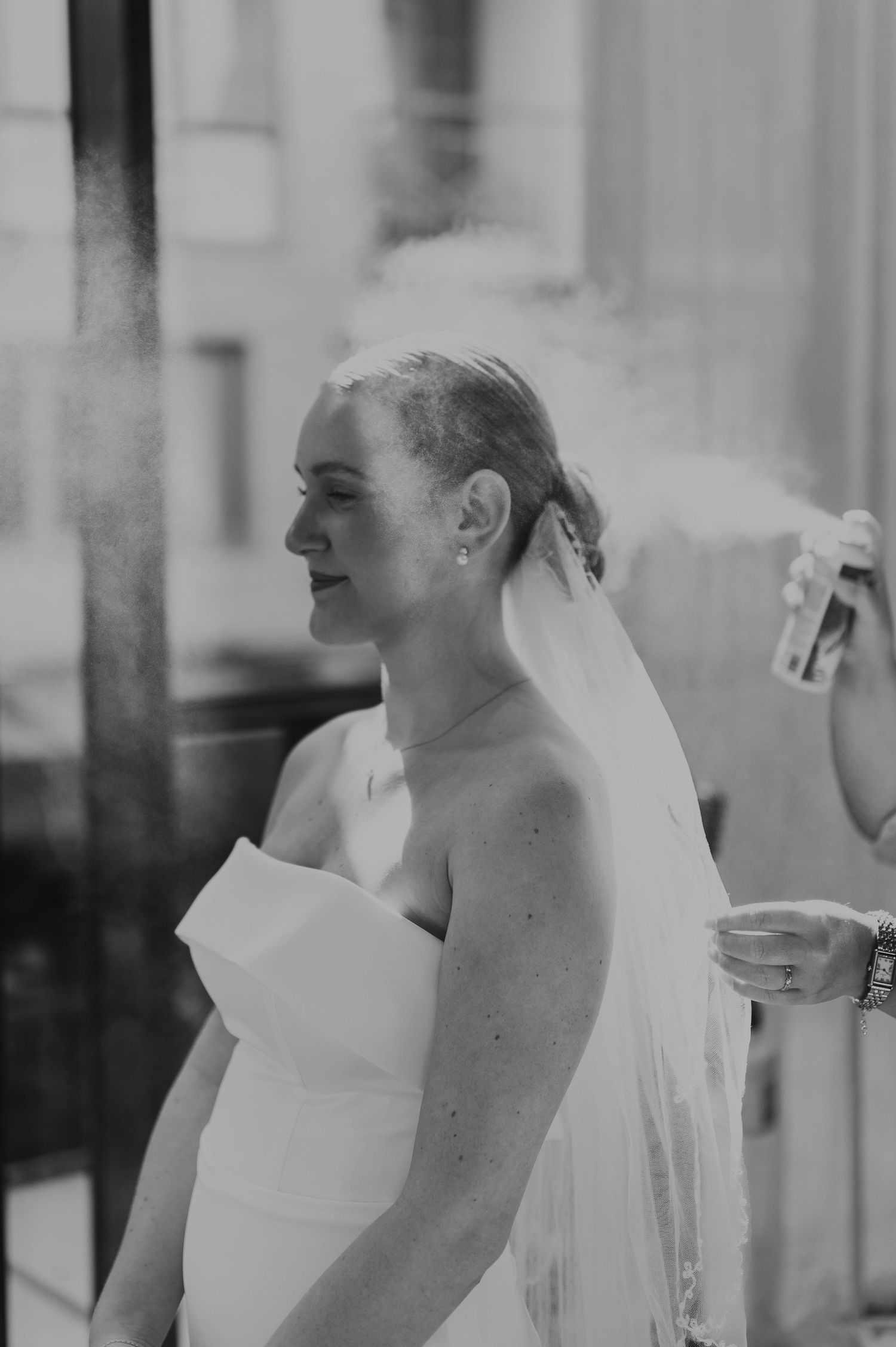 Black and white photo of bride being spritzed with hairspray while wearing a strapless gown.