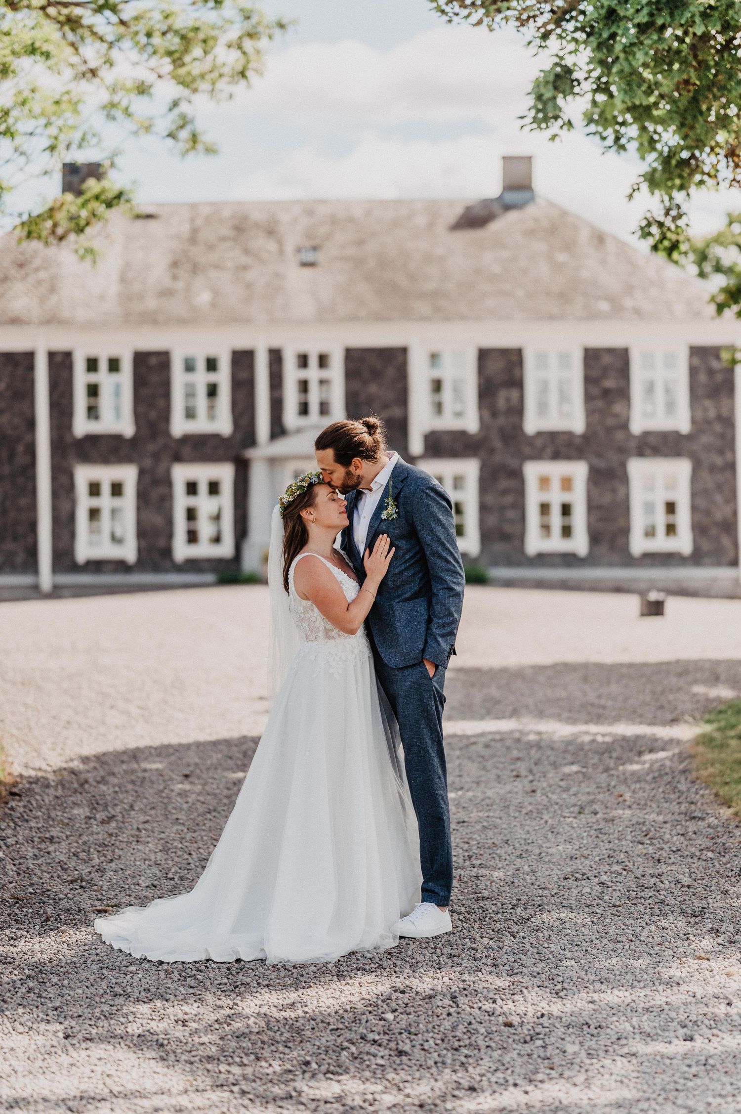 Bride in floral crown and groom in navy suit embracing before a grand historic manor.