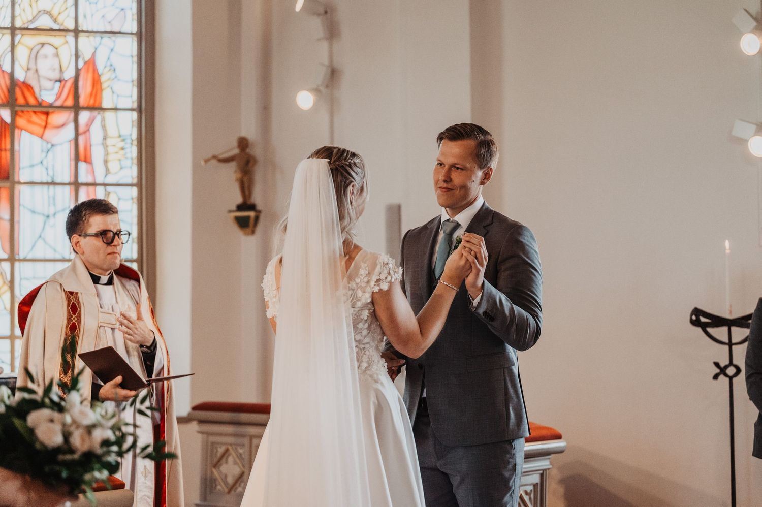 Groom in gray suit holds brides hands during wedding ceremony as officiant reads in a sunlit church.