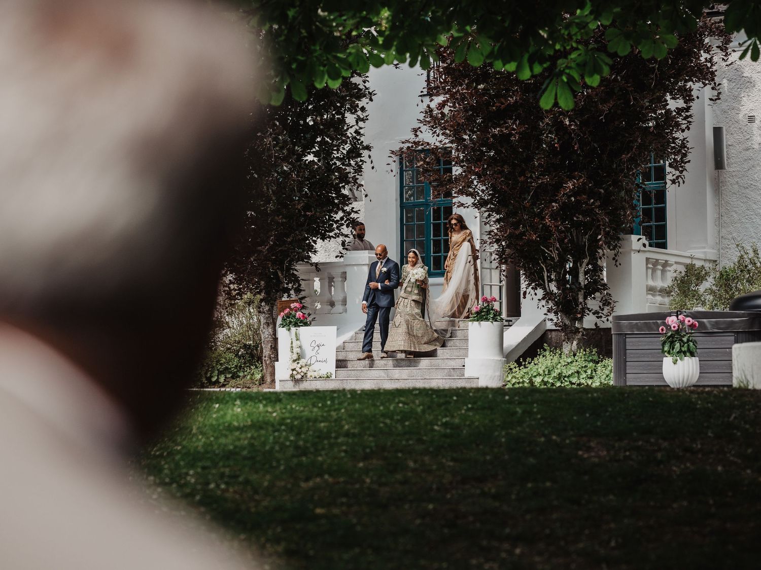 Bride and groom walk toward garden wedding venue framed by lush trees and elegant white columns.