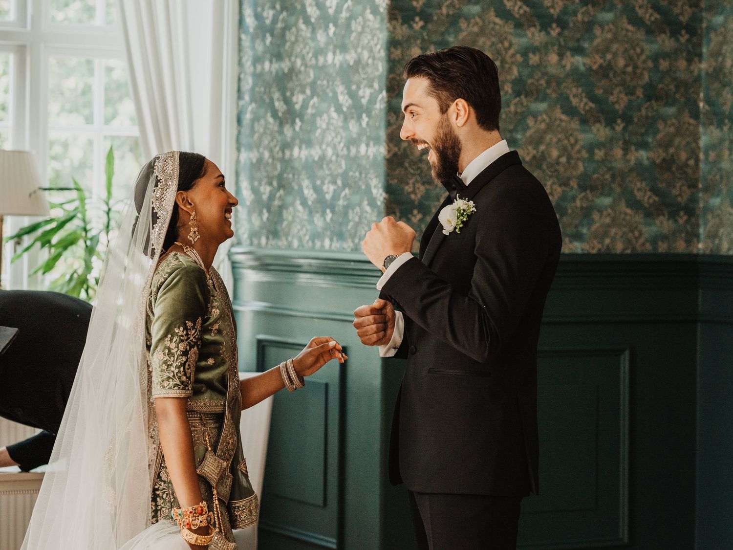 Bride in green embroidered gown with veil and groom in black tuxedo share a joyful moment at elegant wedding.