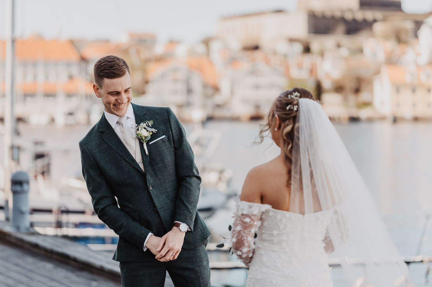 Smiling groom in dark suit faces bride in lace wedding dress at a picturesque harbor venue.