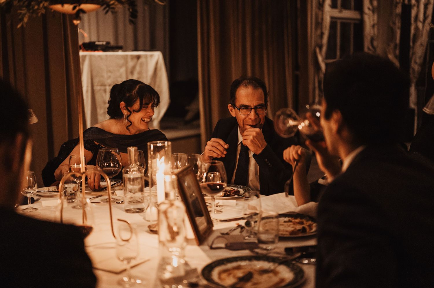 Guests enjoying a candlelit wedding dinner, sharing conversation over wine in an elegant, dimly lit venue.