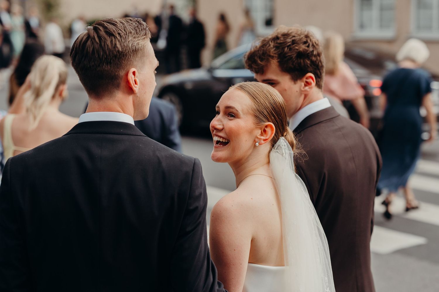 Smiling bride in white veil laughing joyfully with guests outside wedding venue on sunny day.