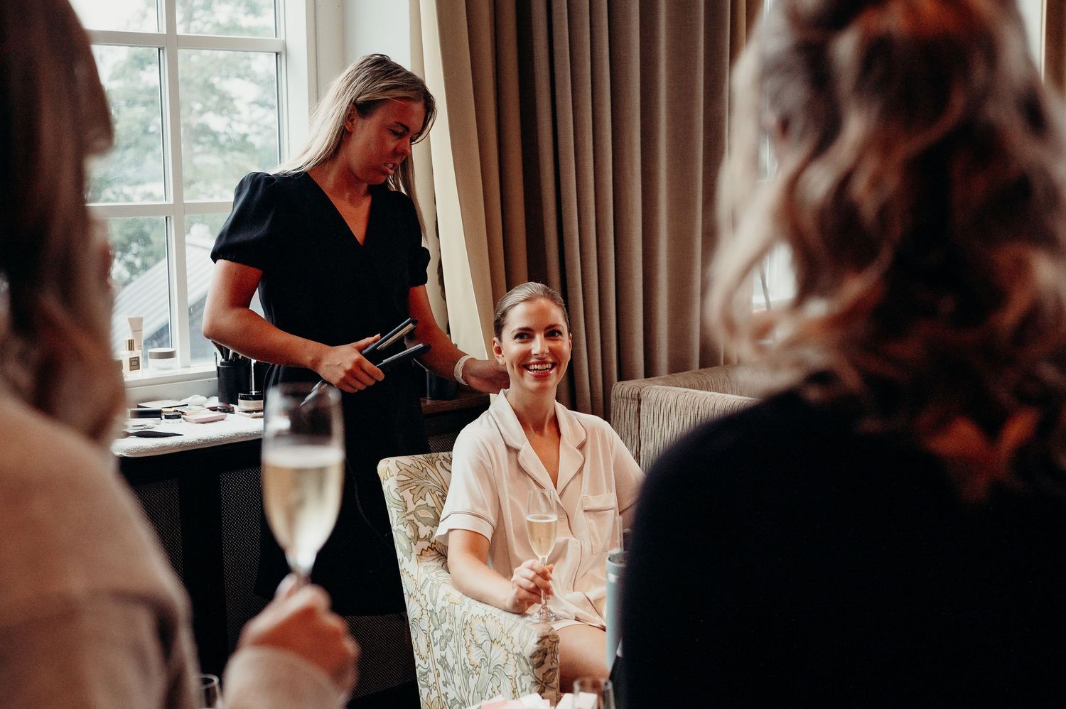 Bridesmaids celebrating with champagne as a woman in black dress pours drinks in an elegant hotel room.