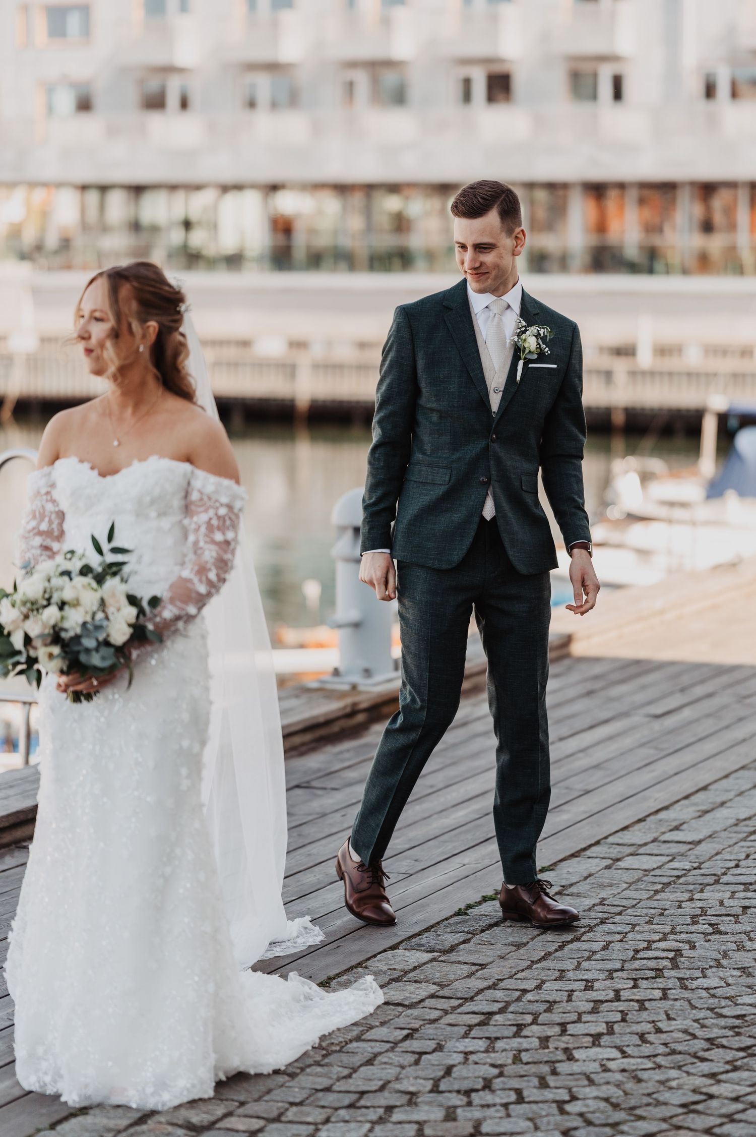 Groom in dark suit walks toward bride holding white bouquet on a cobblestone waterfront dock.