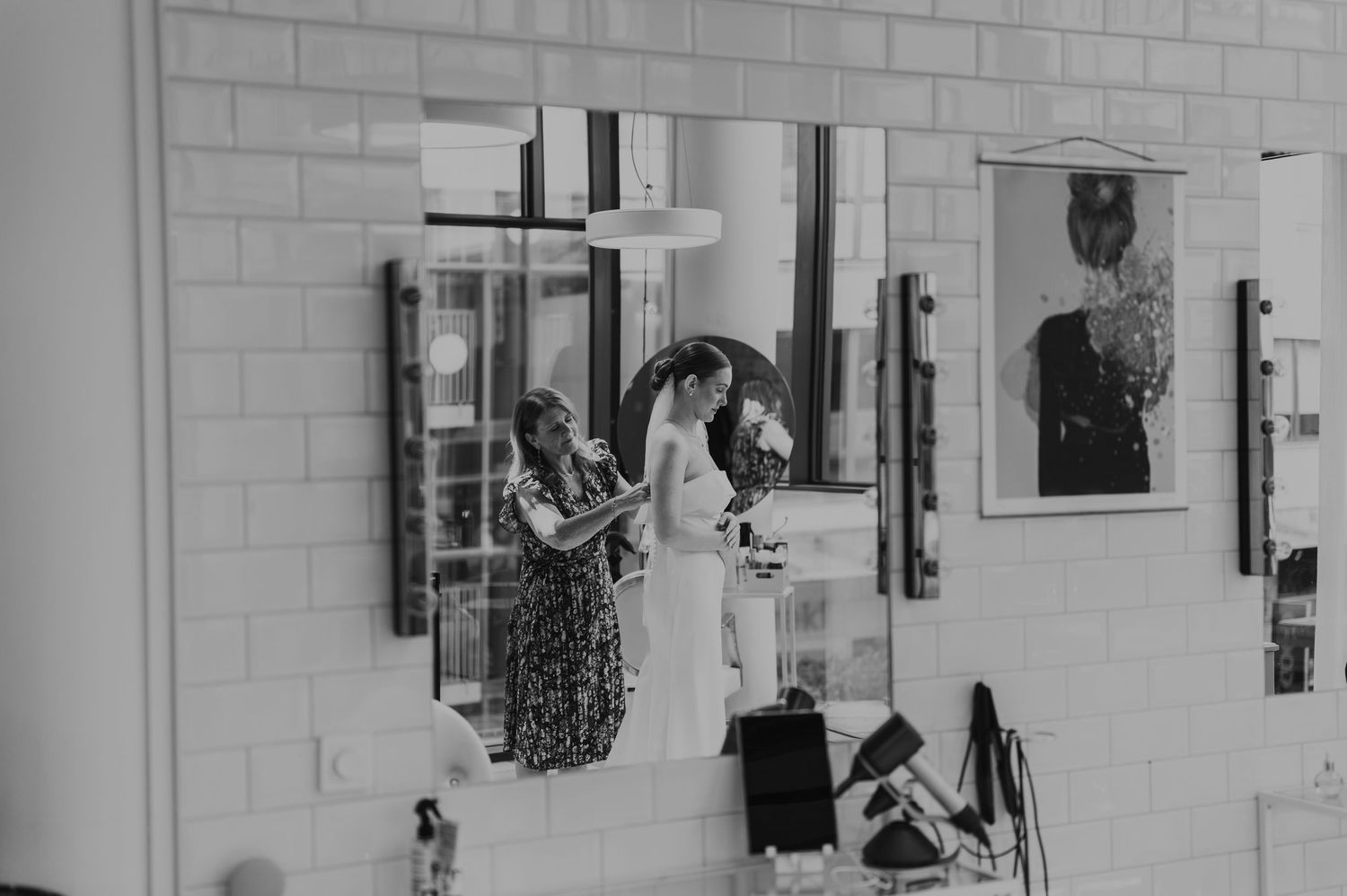 Black and white photo of bride getting ready with hairstylist assistance reflected in mirror.