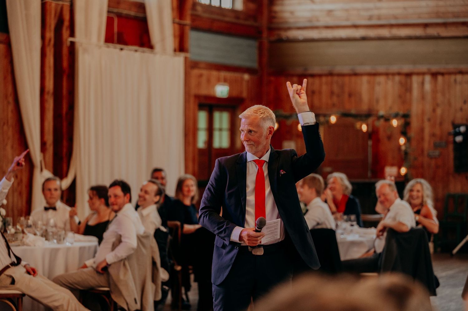 Man in navy suit raising hand while giving speech at elegant wedding reception.