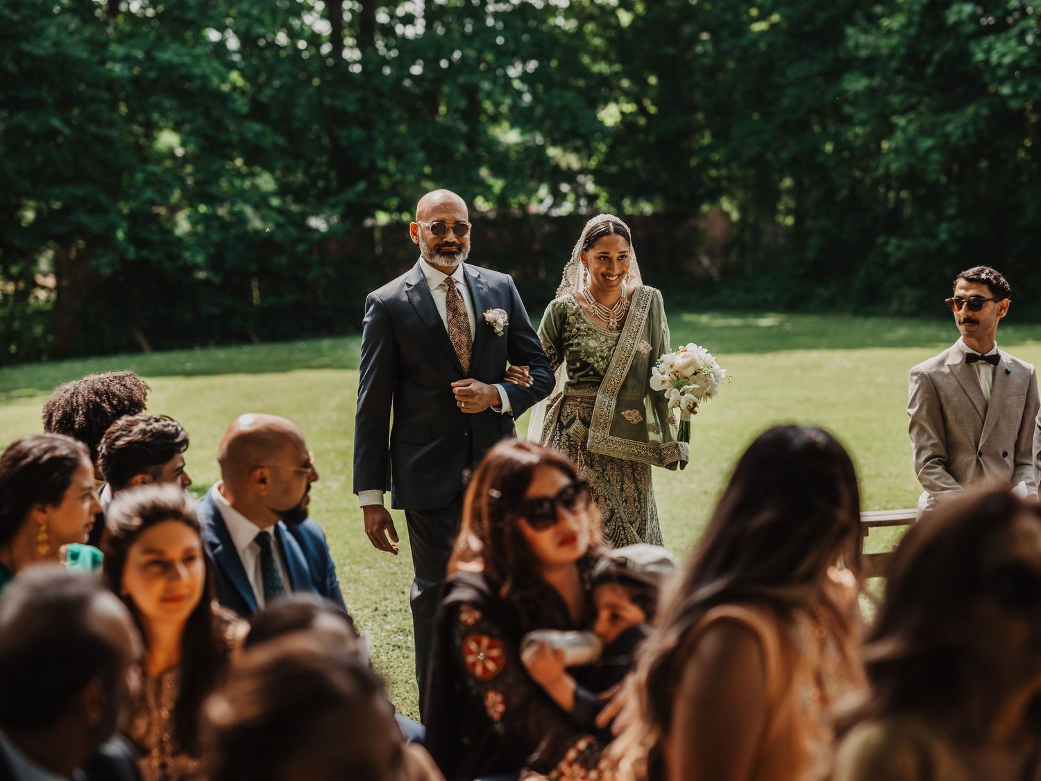 Bride walks down outdoor aisle with escort as wedding guests watch during garden ceremony.